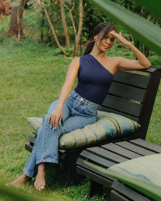 A woman sitting on a bench wearing a one shoulder navy bodysuit and blue jeans.