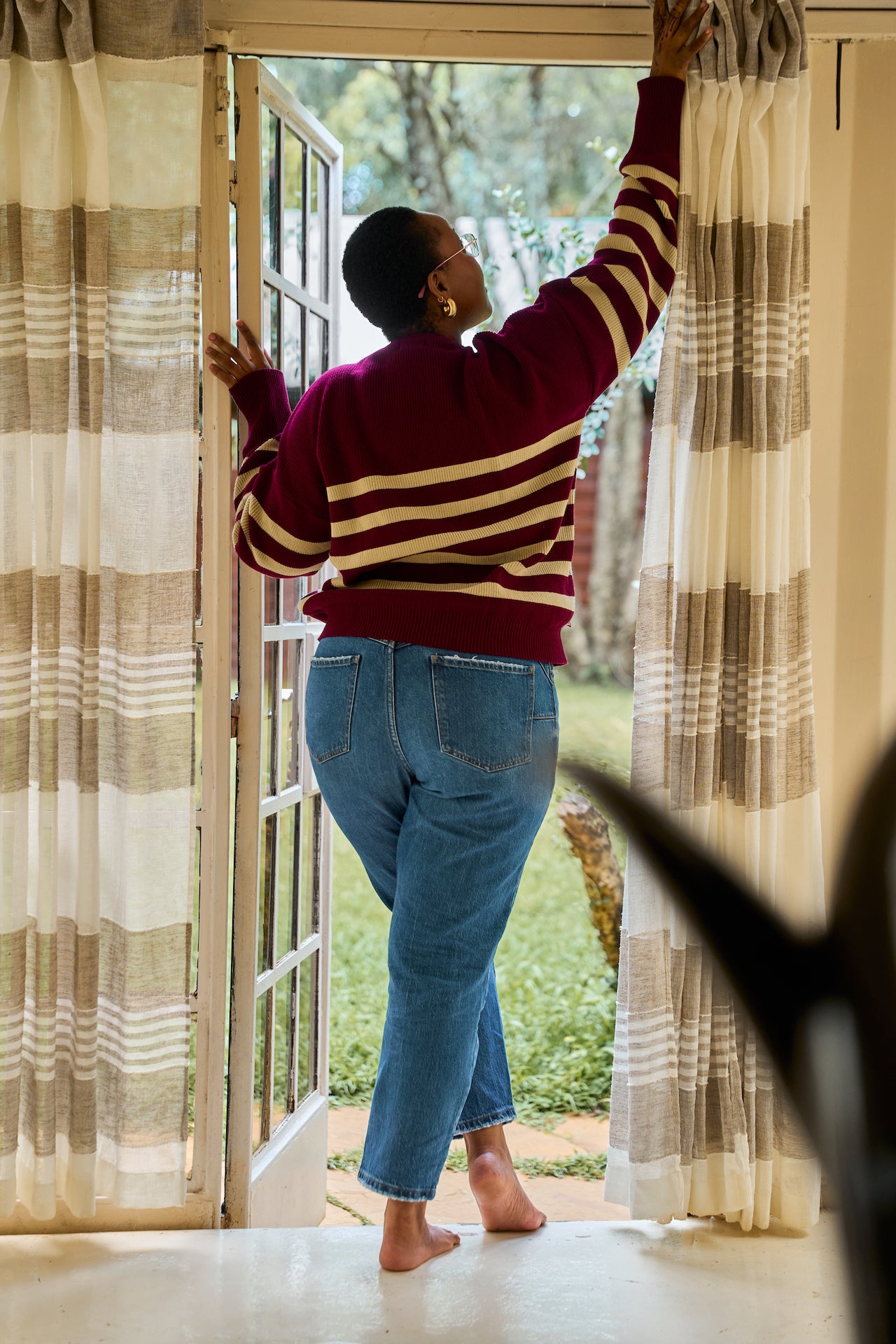 Person in a striped knit sweater and jeans standing by a glass door with curtains.