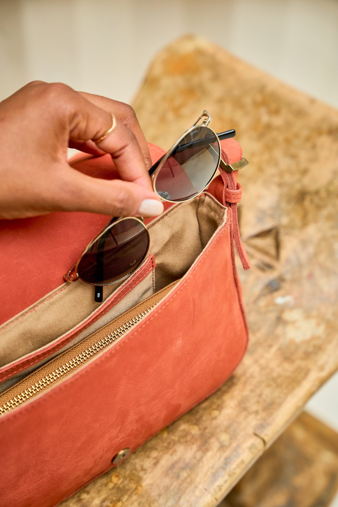 Person placing sunglasses into a red  suede leather handbag on a wooden surface