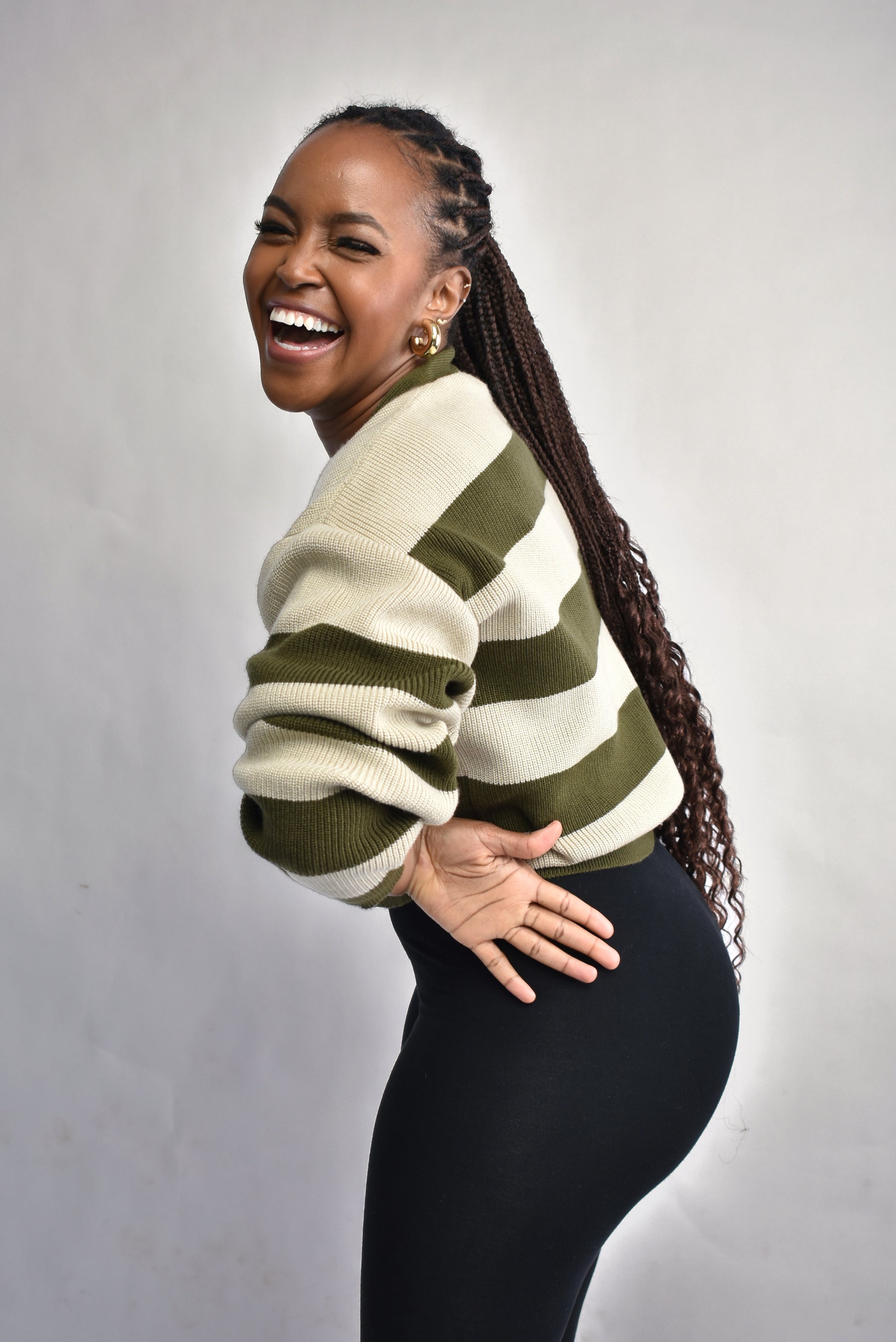 Woman wearing a green and beige striped sweater paired wit black leggings against a gray background