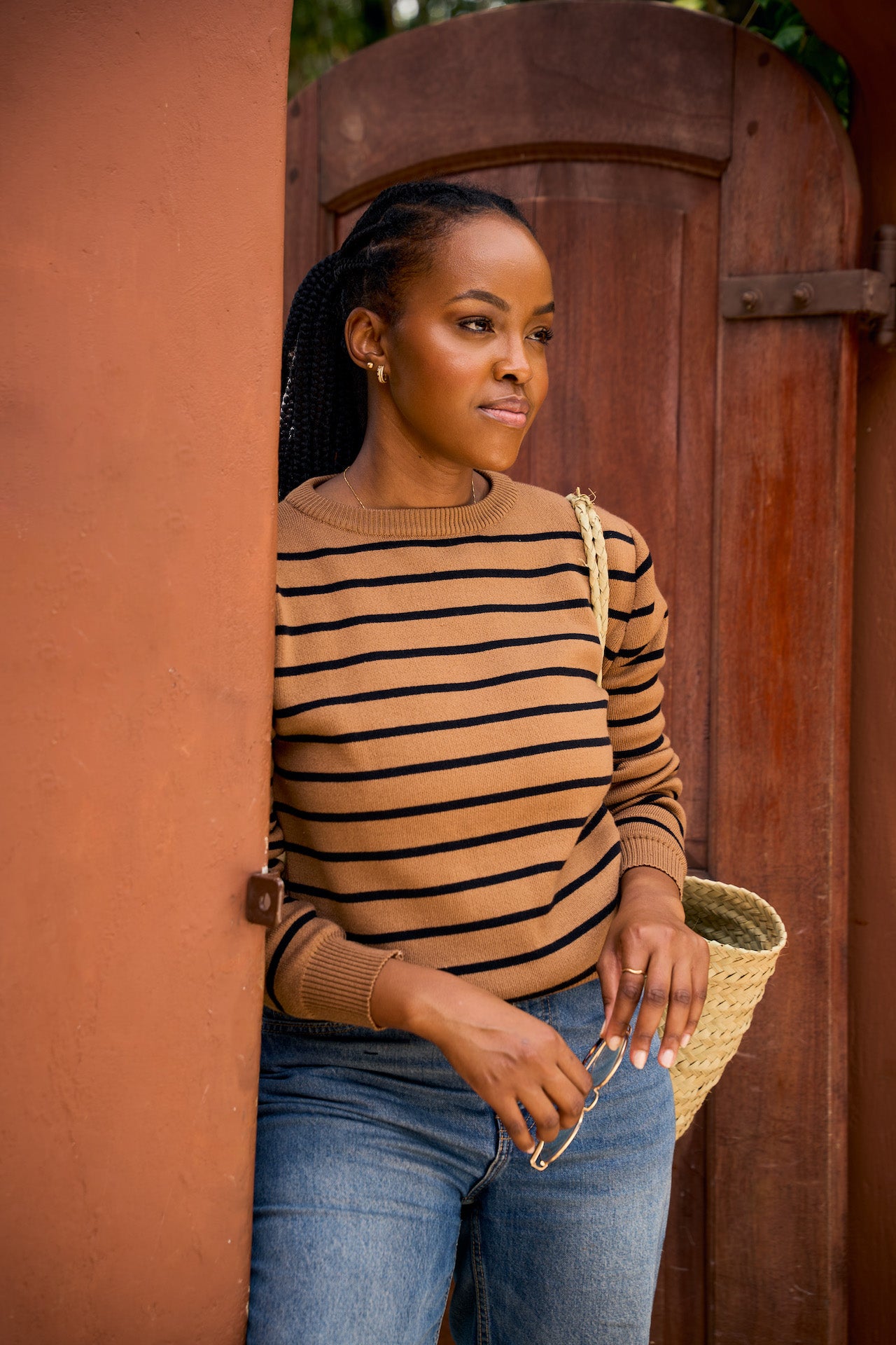 Woman wearing a striped knit sweater and jeans, holding a straw bag, standing against a wooden door.