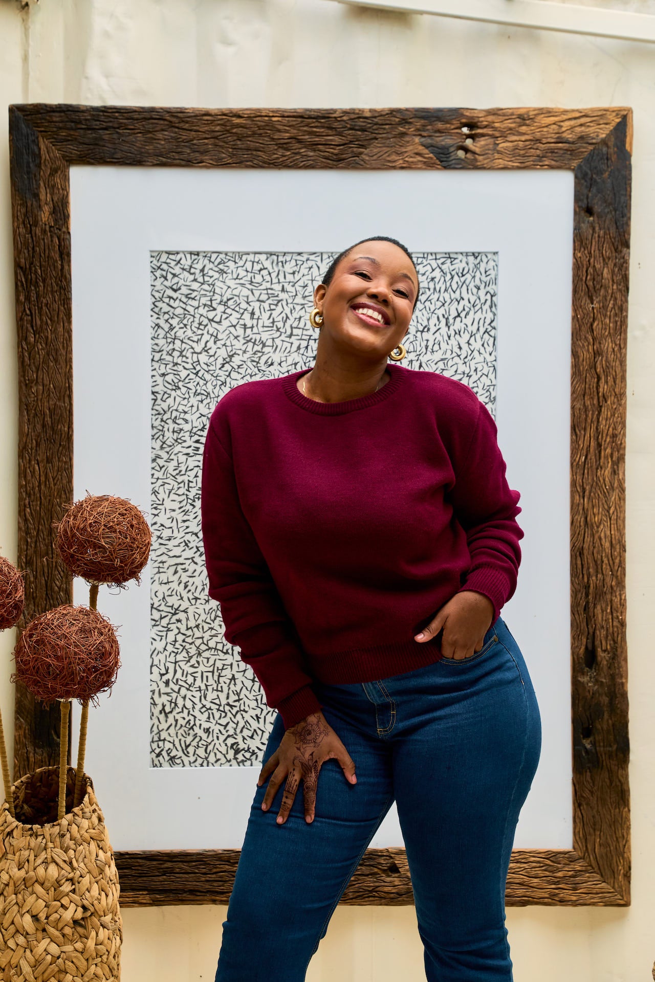 Woman in a maroon shani knit  sweater and blue jeans standing in front of a framed artwork.