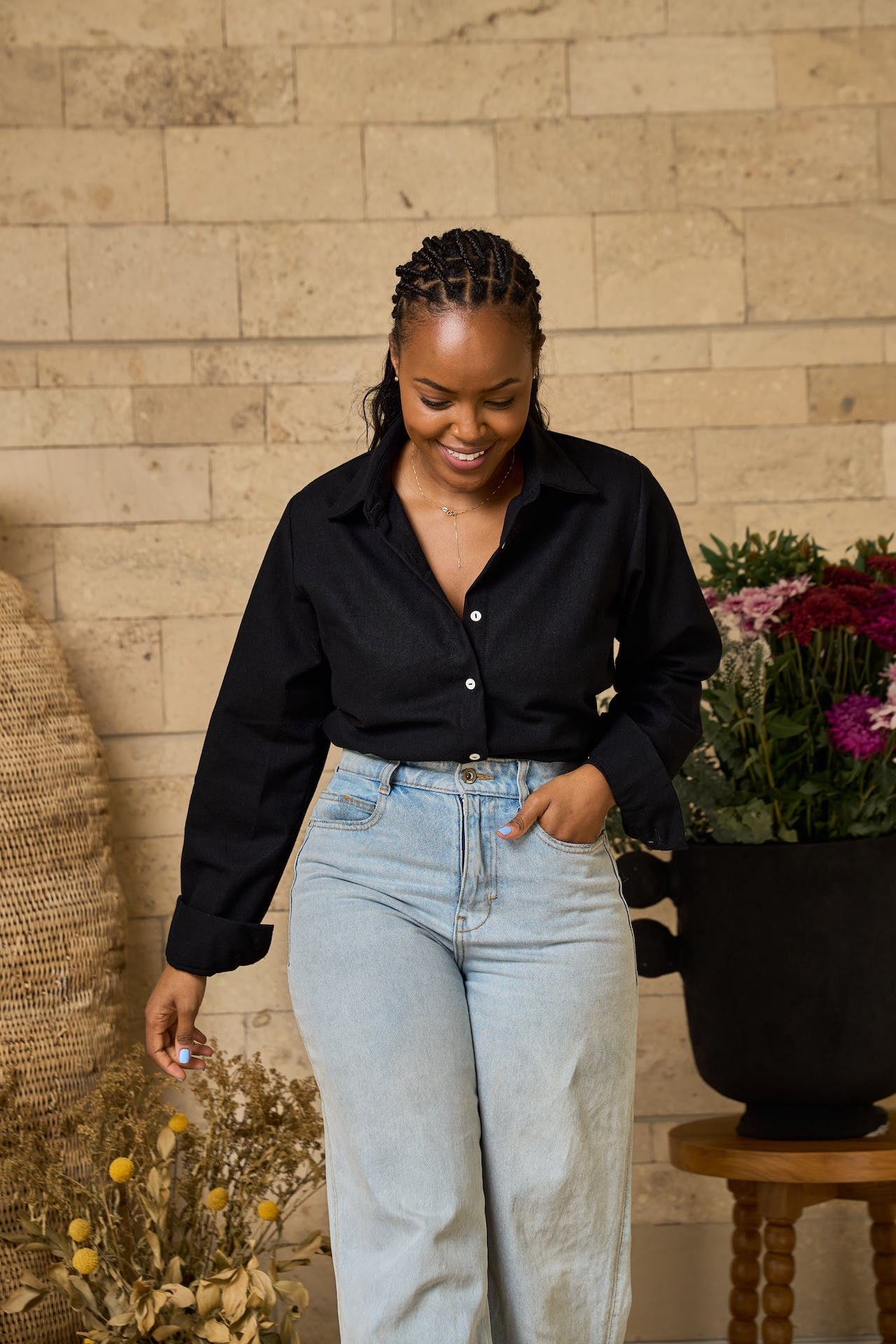 Woman in black linen shirt and light blue jeans standing next to a potted plant against a brick wall.