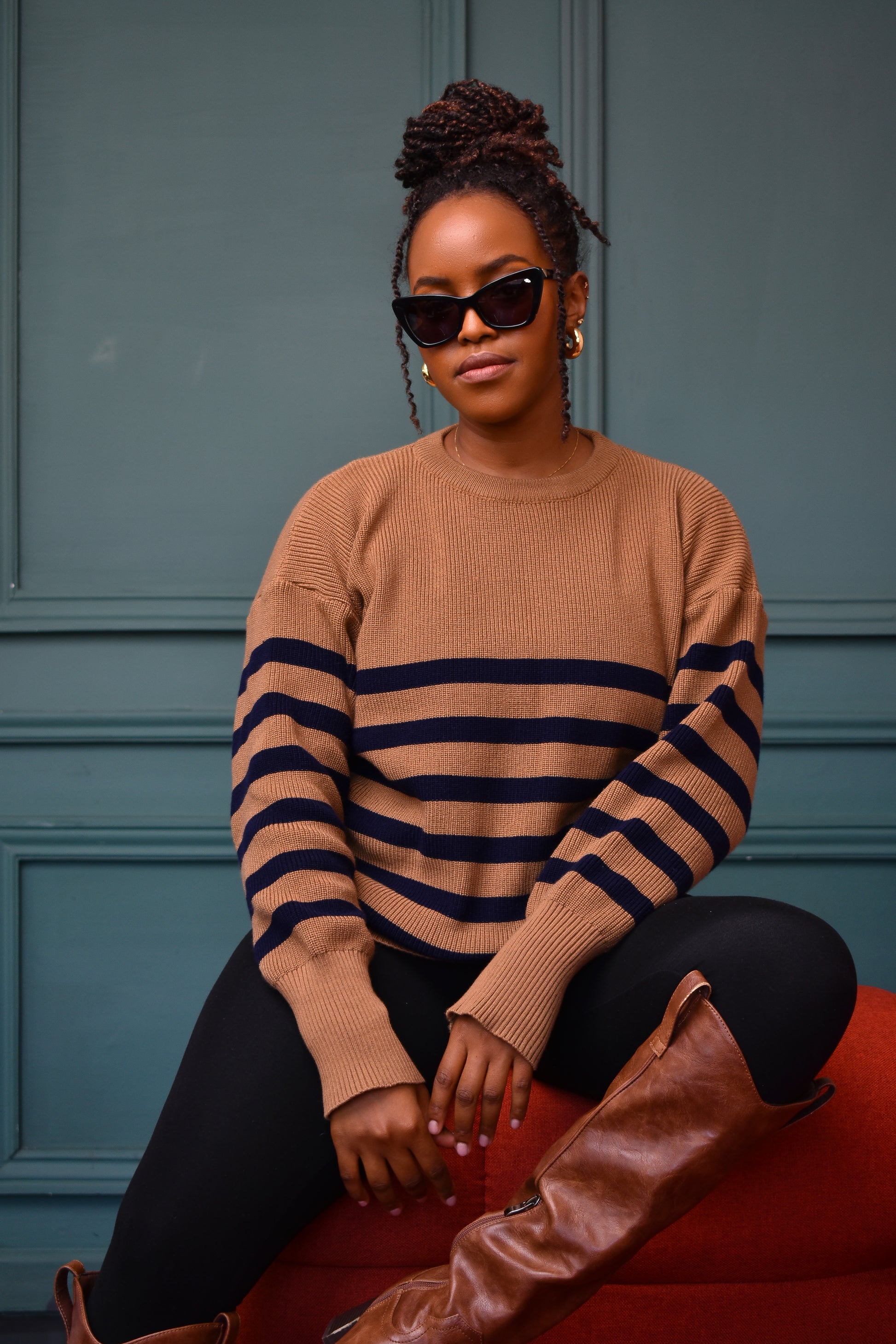 A woman sitting on a red bench against a blue wall, wearing the classic striped knit sweater in a beige and navy color combination, paired with black pants and brown boots, accessorized with sunglasses and earrings.